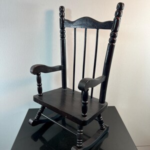 May include: A small, black wooden rocking chair with a curved back and armrests. The chair has a classic design with rounded spindles and rockers. The chair is likely a decorative item or a child's toy.