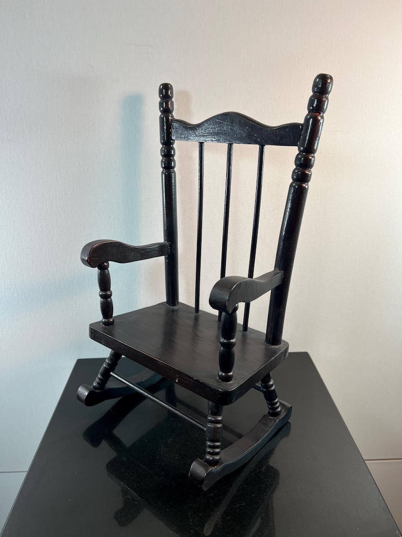 May include: A small, black wooden rocking chair with a curved back and armrests. The chair has a classic design with rounded spindles and rockers. The chair is likely a decorative item or a child's toy.
