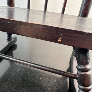 May include: Close-up of a dark brown wooden chair with a spindle back. The chair has a rectangular seat and turned legs. The wood has a glossy finish and shows signs of wear. The chair is likely an antique or vintage piece.