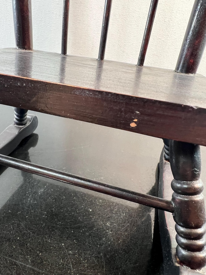 May include: Close-up of a dark brown wooden chair with a spindle back. The chair has a rectangular seat and turned legs. The wood has a glossy finish and shows signs of wear. The chair is likely an antique or vintage piece.