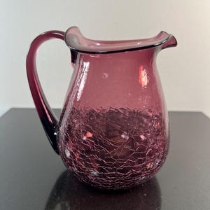 May include: A plum-colored glass pitcher with a crackle finish. The pitcher has a curved handle and a ruffled rim. The glass is translucent, allowing light to pass through. The pitcher is likely used for serving beverages.