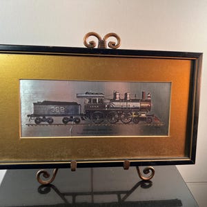 May include: Framed vintage train print. The artwork features a detailed illustration of a steam locomotive and coal car, set against a silver background. The frame is black with a gold inner border. The print is displayed on a decorative metal stand.