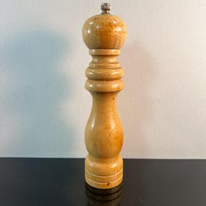 May include: A wooden pepper mill with a light brown finish. The mill has a bulbous top, a cylindrical body with decorative rings, and a metal cap. It stands upright on a black surface against a white background.