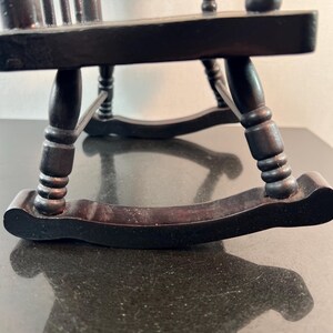 May include: A miniature black wooden rocking chair. The chair has a slatted back and curved rockers. The legs are turned and the chair appears to be made of dark stained wood. The chair is sitting on a dark surface.
