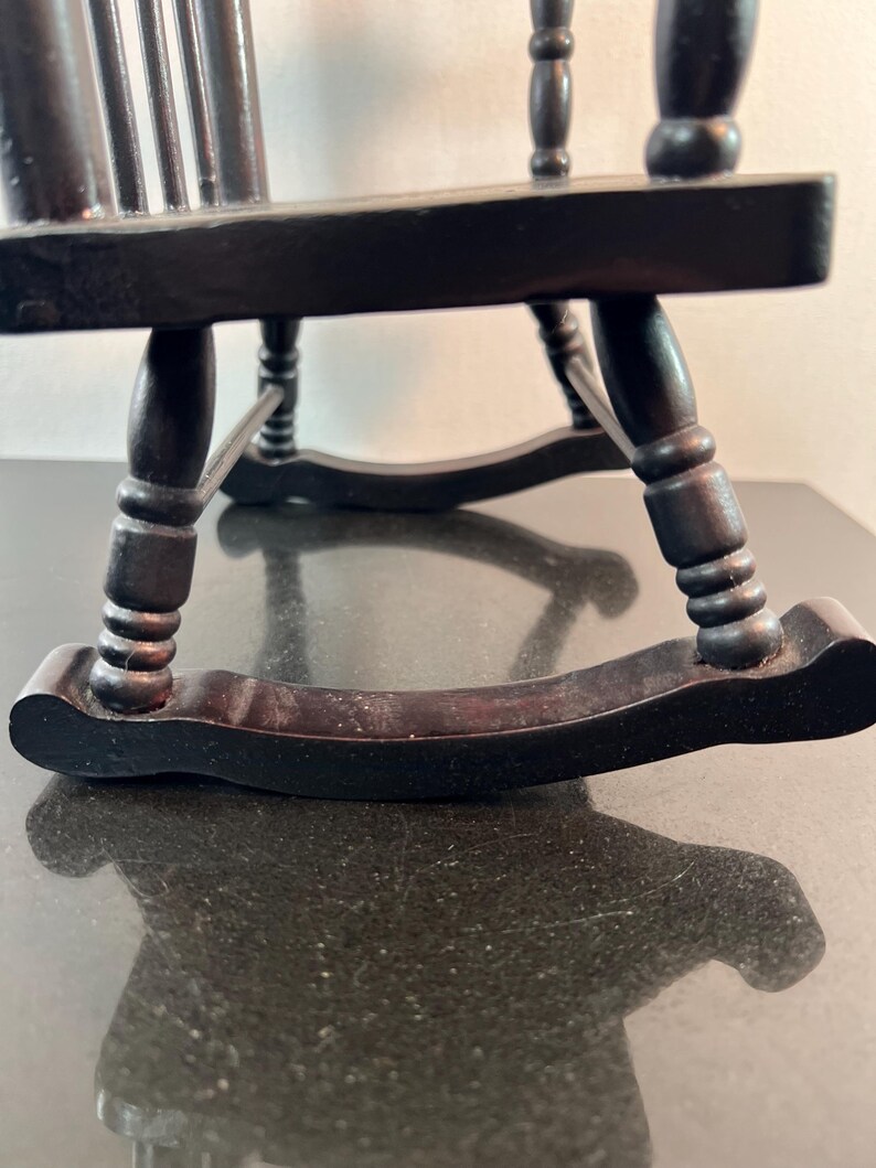May include: A miniature black wooden rocking chair. The chair has a slatted back and curved rockers. The legs are turned and the chair appears to be made of dark stained wood. The chair is sitting on a dark surface.