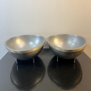May include: Two silver bowls with a gold rim and three legs each. The bowls are sitting on a black surface.