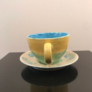 May include: A yellow and blue ceramic teacup and saucer set with a green band around the cup. The saucer has a blue and white pattern.