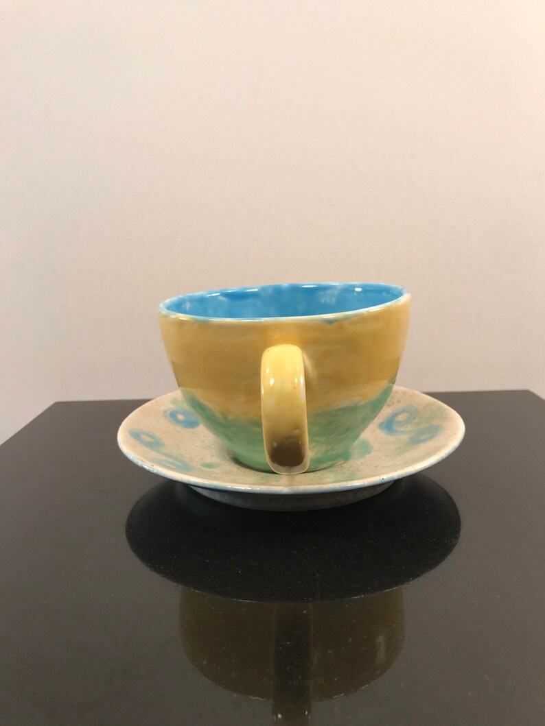 May include: A yellow and blue ceramic teacup and saucer set with a green band around the cup. The saucer has a blue and white pattern.