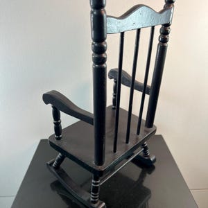May include: A small, black wooden rocking chair with a slatted back and curved rockers. The chair has armrests and a classic design, perfect for a doll or decorative display. The chair is painted black.