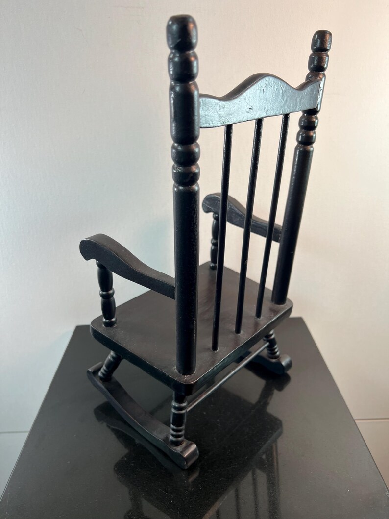 May include: A small, black wooden rocking chair with a slatted back and curved rockers. The chair has armrests and a classic design, perfect for a doll or decorative display. The chair is painted black.