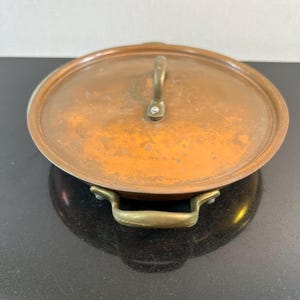 May include: Oval copper pot with a matching lid. The pot has a brass handle on the lid and two brass handles on the sides. The copper has a warm, aged patina. A classic kitchenware item.