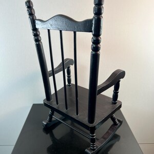 May include: A small, black wooden rocking chair. The chair has a slatted back, curved arms, and a seat. The chair is designed with a classic, traditional style. The chair is made of wood and has a glossy finish.
