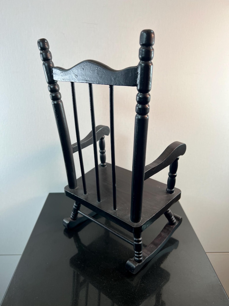 May include: A small, black wooden rocking chair. The chair has a slatted back, curved arms, and a seat. The chair is designed with a classic, traditional style. The chair is made of wood and has a glossy finish.