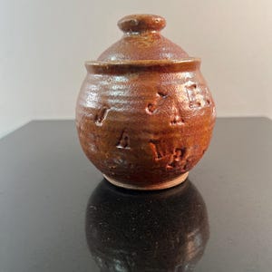 May include: A small, round, brown ceramic jar with a lid. The jar has a textured surface and the word "Jars" is carved into the side. The lid has a small knob on top. The jar is sitting on a black surface.