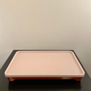 May include: A pink plastic rectangular storage container with a brown base. The container has a slightly raised rim and a small circle in the center of the lid.