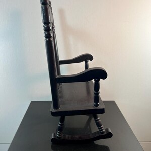 May include: A small, black wooden rocking chair. The chair has a slatted back, curved rockers, and armrests. The chair is made of wood and has a glossy finish. The chair is a miniature version of a traditional rocking chair.