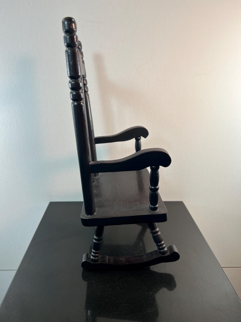 May include: A small, black wooden rocking chair. The chair has a slatted back, curved rockers, and armrests. The chair is made of wood and has a glossy finish. The chair is a miniature version of a traditional rocking chair.