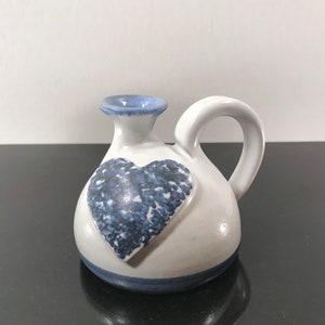 May include: A small white ceramic pitcher with a blue heart design. The pitcher has a handle and a spout. The heart is raised and has a blue and white speckled pattern.