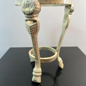 May include: A small, antique-style, metal stand with a round top and three ornate legs. The legs are detailed with shell and scroll designs. The stand has a circular support ring near the base and is finished in a distressed, aged gold color.