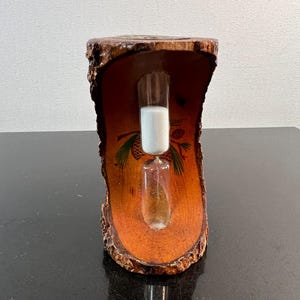 May include: A decorative hourglass with white sand inside a clear glass tube. The hourglass is set in a wooden frame with a natural bark edge and a painted pinecone design. The wood has a warm, orange-brown hue.