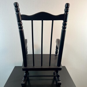 May include: A small, black wooden rocking chair. The chair has a curved back with vertical slats and rounded armrests. The legs are curved, and the chair is painted in a glossy black finish. The chair is designed for decorative purposes.