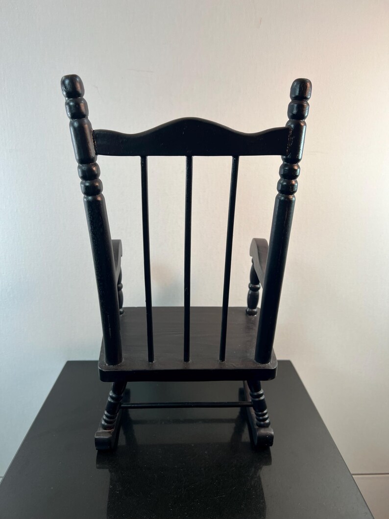 May include: A small, black wooden rocking chair. The chair has a curved back with vertical slats and rounded armrests. The legs are curved, and the chair is painted in a glossy black finish. The chair is designed for decorative purposes.