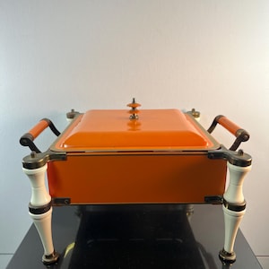 May include: An orange rectangular serving dish with a lid, brass trim, and handles. The dish sits on four white and brass legs. The handles are brown. The dish is likely used for keeping food warm.