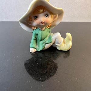 May include: A ceramic figurine of a sitting elf wearing a green shirt and a white hat. The elf has blonde hair, brown eyes, and a smiling face.