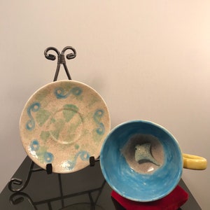 May include: A blue and white ceramic teacup and saucer with a green and white speckled glaze. The teacup has a small, white ceramic stingray inside. The saucer is on a black metal easel.