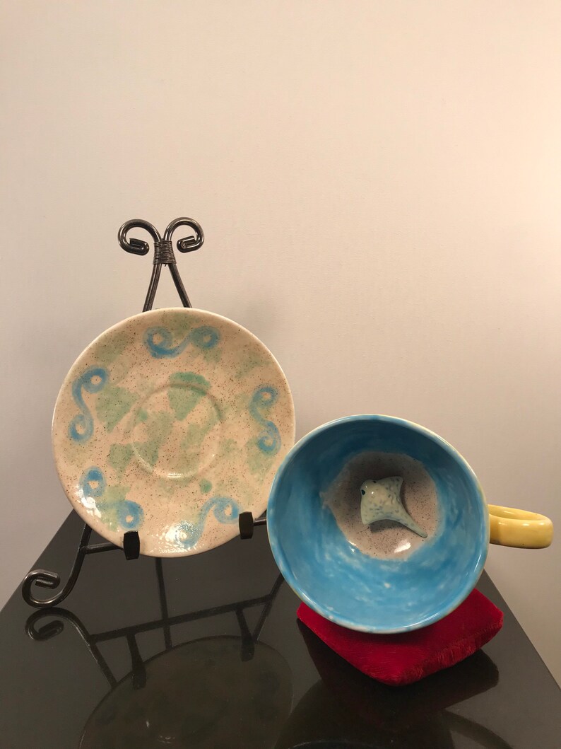 May include: A blue and white ceramic teacup and saucer with a green and white speckled glaze. The teacup has a small, white ceramic stingray inside. The saucer is on a black metal easel.