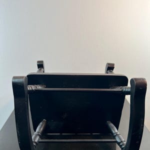 May include: A small, black wooden chair with a rectangular seat and curved back supports. The chair has turned legs and a horizontal bar for support. The chair is positioned against a white wall.