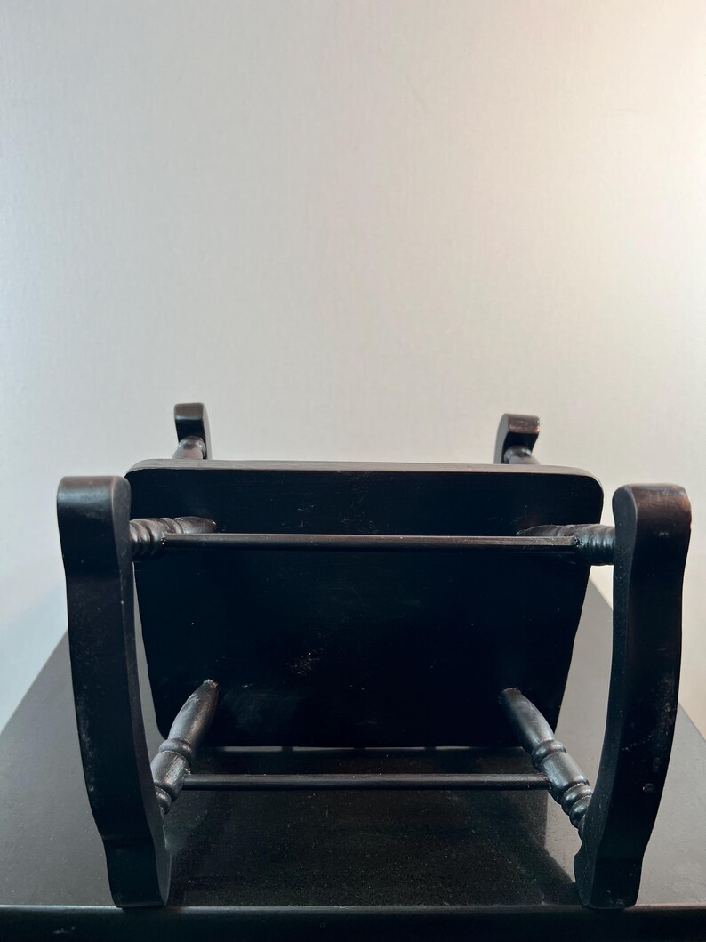 May include: A small, black wooden chair with a rectangular seat and curved back supports. The chair has turned legs and a horizontal bar for support. The chair is positioned against a white wall.
