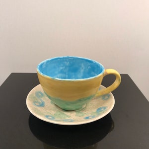 May include: A yellow and blue ceramic teacup and saucer set with a light blue interior and a white speckled design.