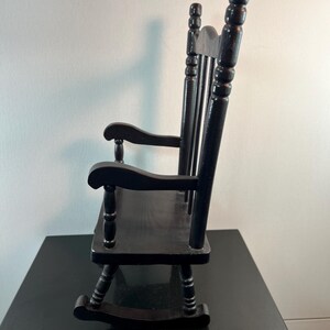 May include: A small, black wooden rocking chair. The chair has a slatted seat, curved rockers, and a high back with three vertical spindles. The armrests are curved and supported by turned posts. The chair is designed for a doll or small child.