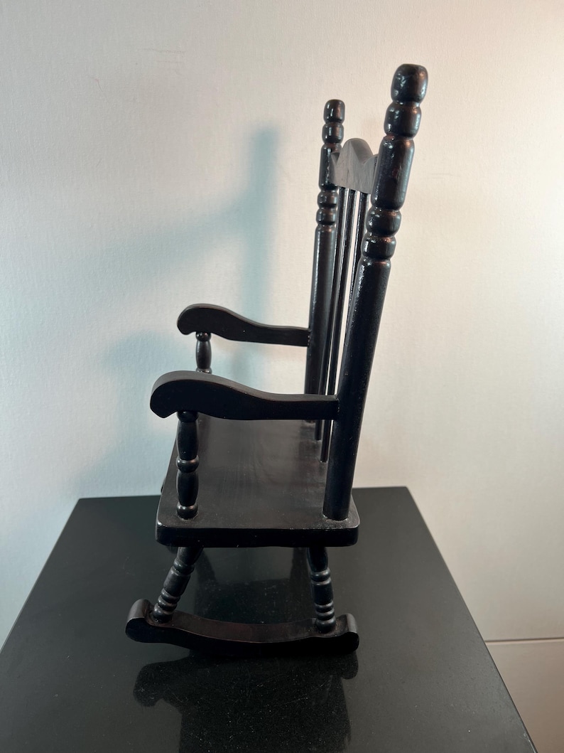 May include: A small, black wooden rocking chair. The chair has a slatted seat, curved rockers, and a high back with three vertical spindles. The armrests are curved and supported by turned posts. The chair is designed for a doll or small child.