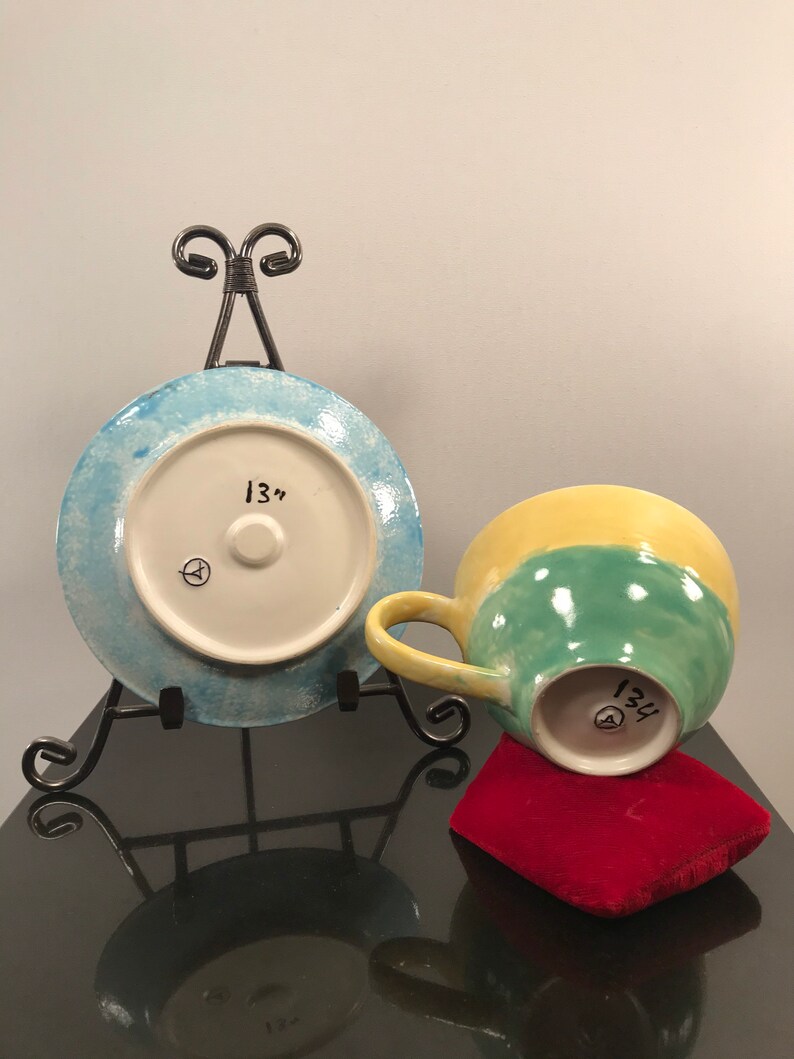 May include: A blue and white ceramic plate with a 13" diameter, and a yellow, green, and white ceramic mug with a 13" diameter. Both pieces are on a black surface.