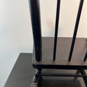 May include: Black wooden chair with a spindle back. The chair has a dark finish and a simple design. The chair is on a black surface. The chair is a decorative item.