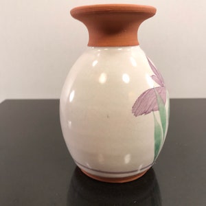 May include: A small, white ceramic vase with a brown terracotta rim and base. The vase is decorated with a purple flower and green leaves.