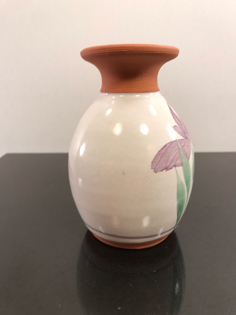 May include: A small, white ceramic vase with a brown terracotta rim and base. The vase is decorated with a purple flower and green leaves.