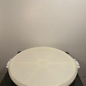 May include: A round, off-white plastic container with a lid. The lid has a slightly raised center and two white handles on opposite sides. The container is sitting on a black surface.