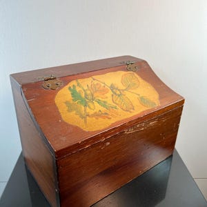 May include: A wooden box with a hinged lid and brass hardware. The lid features a painted design of acorns and leaves in shades of green, orange, and yellow against a gold background. The box is stained a dark brown color.