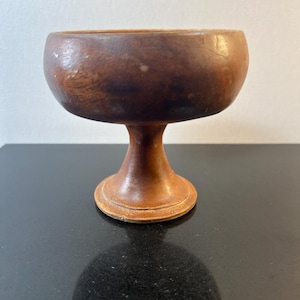 May include: A small, vintage wooden chalice with a rounded bowl and a pedestal base. The chalice is made of a warm brown wood with visible grain and a smooth finish. The base is circular and the stem is slender.