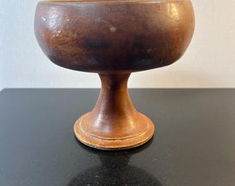 May include: A small, vintage wooden chalice with a rounded bowl and a pedestal base. The chalice is made of a warm brown wood with visible grain and a smooth finish. The base is circular and the stem is slender.