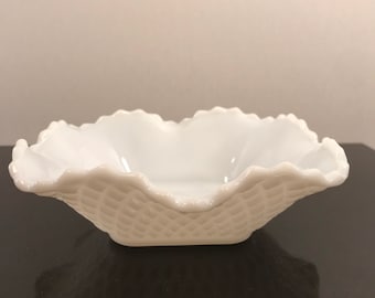 May include: A white, vintage, ruffled-edge bowl with a basketweave pattern. The bowl has a square base and a scalloped rim. The bowl is made of opaque white glass and is likely used for serving.