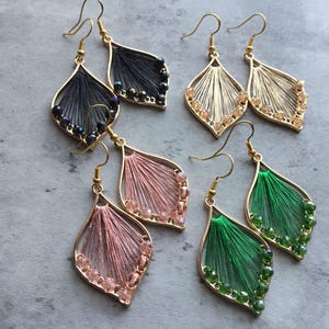 以下が含まれることがあります： 金色の金属製の留め具とカラフルな糸のアクセントが付いた、4ペアのドロップイヤリング。各ペアは、黒、ベージュ、ピンク、緑の異なる糸の色をしています。