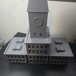 May include: A gray, 3D-printed model of a building complex. The model features a central clock tower with a clock face and a pitched roof, flanked by lower rectangular structures with multiple windows. The overall design is a miniature architectural representation.
