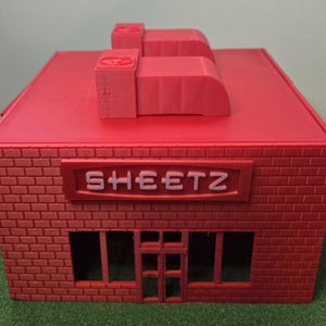 May include: A red brick-textured model of a Sheetz convenience store. The model has a red roof with two rectangular structures and the Sheetz logo above the windows. The model is on a green surface.