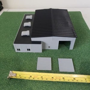 May include: A white model building with a black roof and three air vents. The building has three doors and four windows. The building is sitting on a green surface with two gray rectangular pieces in front of it.