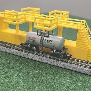 May include: A model train car with a yellow platform and stairs. The car is carrying a gray tank car with the text "DUPONT GATX 62673" and "BUDIUM CAR COATINGS".