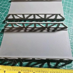 Puede incluir: Tres secciones de puente de plástico gris con un diseño de armadura. Las secciones tienen 10 pulgadas de largo y se miden con una cinta métrica amarilla.